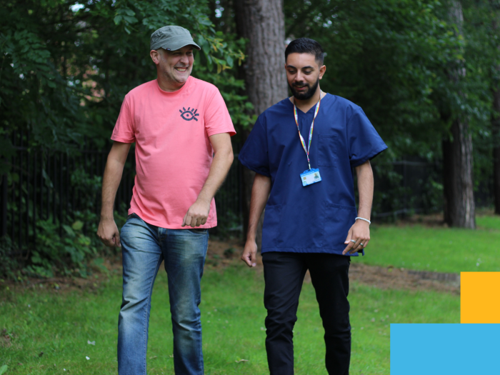 Two people walk in green area by trees, smiling and talking, one wearing casual clothes and the other in medical clothing